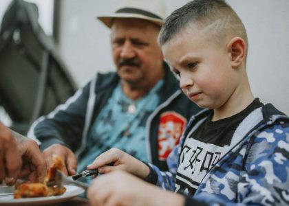 A child and his father share a meal outdoors, enjoying grilled food together.